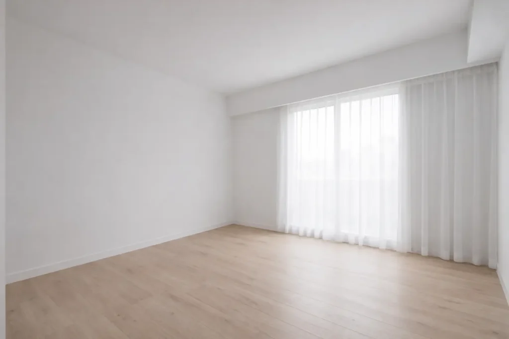 Empty modern bedroom with white walls, light wood flooring, and sheer curtains letting in natural daylight