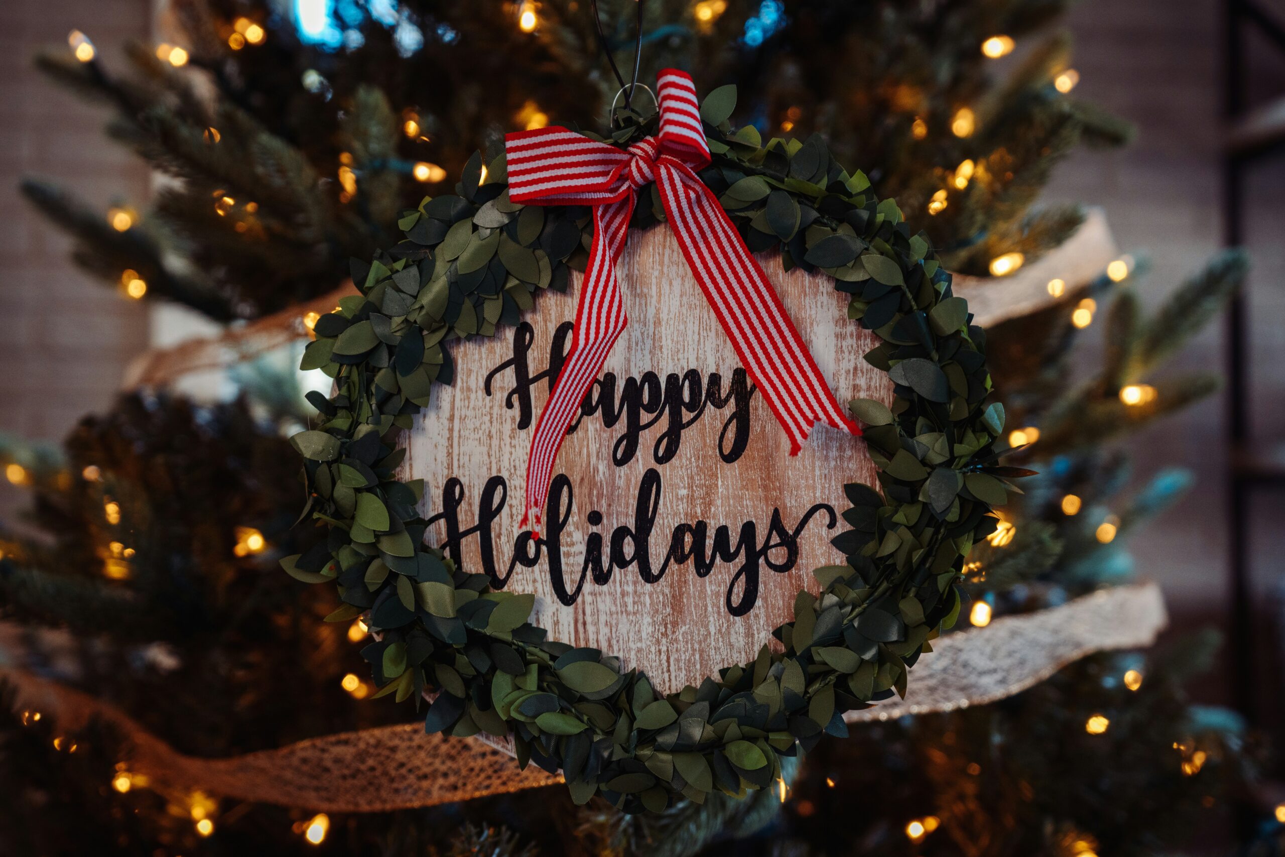 Christmas wreath with greenery leaves and a red ribbon bow hanging in front of warm holiday lights, featuring a rustic “Happy Holidays” wooden sign